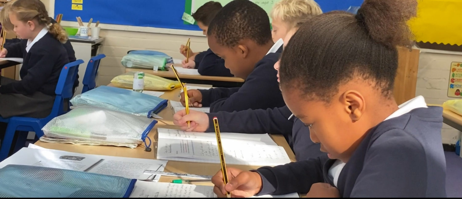 A group of pupils are seen sat at their desks, writing in their exercise books during a lesson.